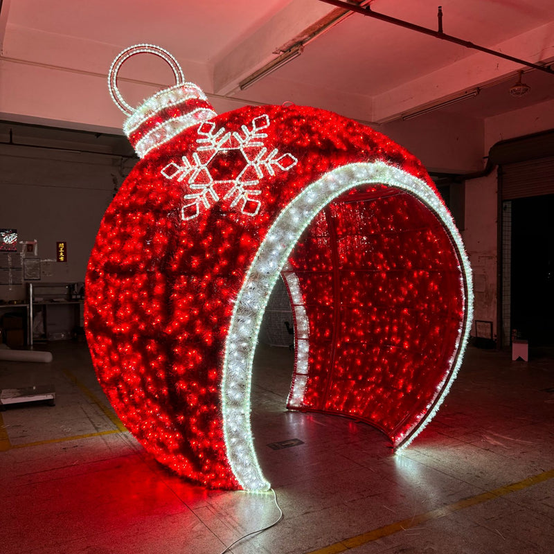 Large illuminated red walk-through Christmas ornament with white snowflake design, displayed in an indoor production facility with glowing LED outline